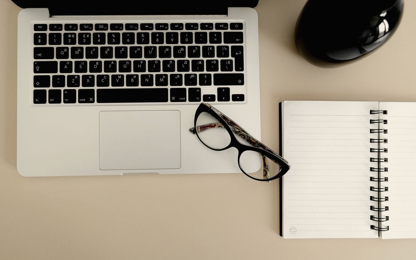 Laptop, Brille und ein aufgeschlagenes Notizbuch mit Stift auf einer sandfarbenen Arbeitsfläche